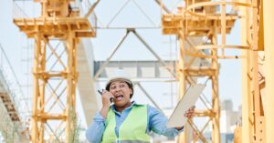 Female engineer using walkie-talkie while holding clipboard on a construction site.